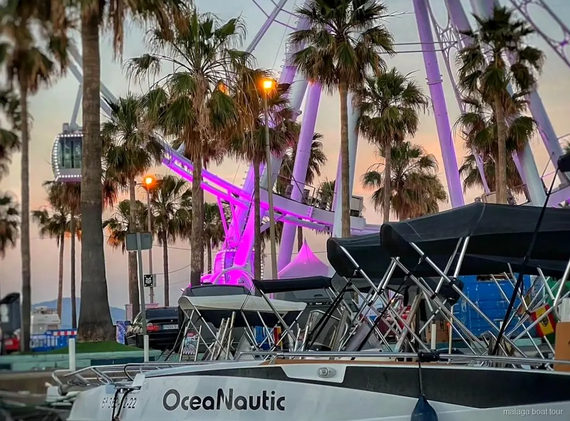 Barcos atracados ao entardecer com palmeiras e roda gigante iluminada antes de um passeio de barco em Málaga