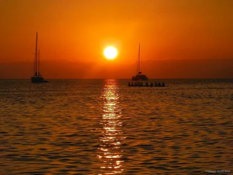Pôr do sol laranja sobre o mar com dois veleiros e um catamarã em silhueta num passeio de barco em Málaga