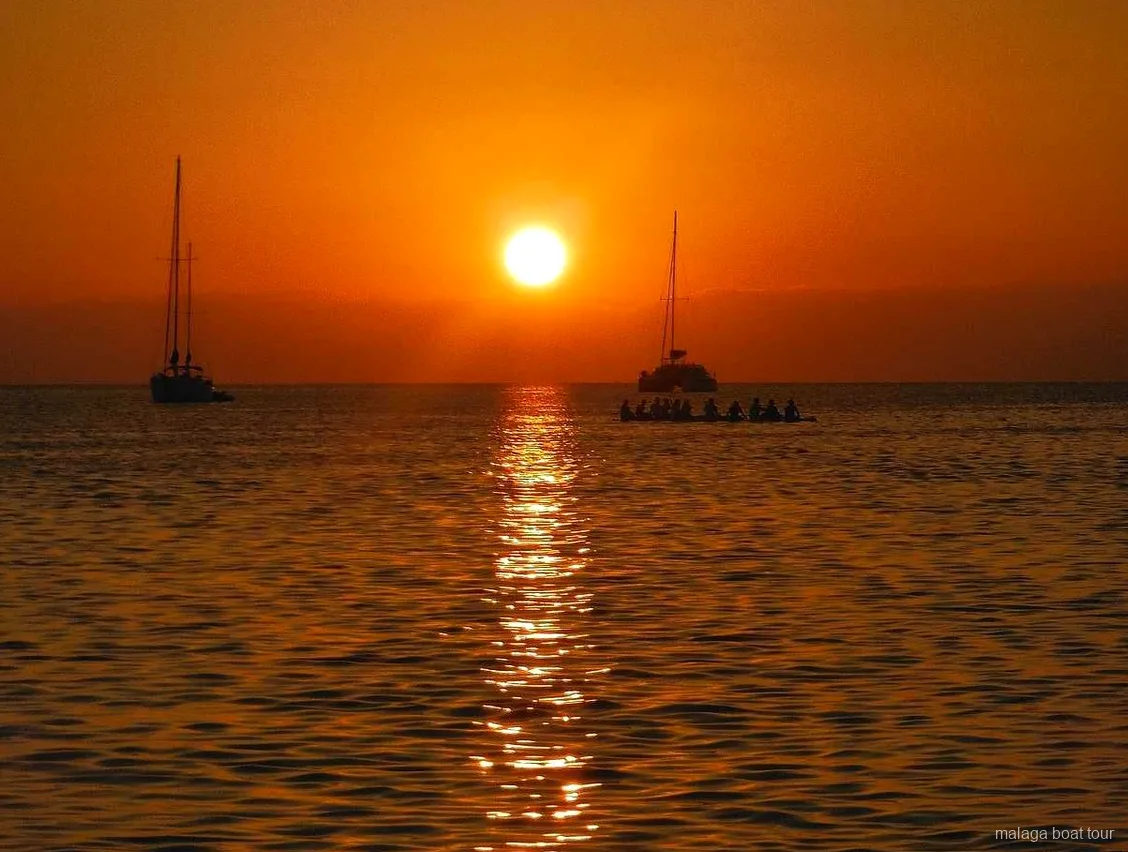Pôr do sol laranja sobre o mar com dois veleiros e um catamarã em silhueta num passeio de barco em Málaga