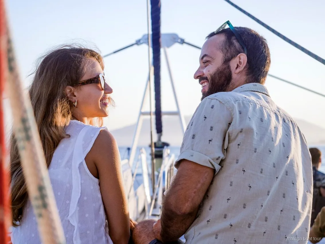 Casal a sorrir no convés de um catamarã com luz quente de fim de tarde num passeio de barco em Málaga