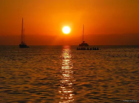 Posta de sol taronja sobre el mar amb dos velers i un catamarà en silueta en una excursió en vaixell a Màlaga