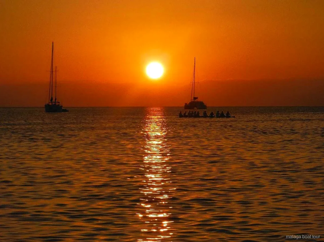 Posta de sol taronja sobre el mar amb dos velers i un catamarà en silueta en una excursió en vaixell a Màlaga
