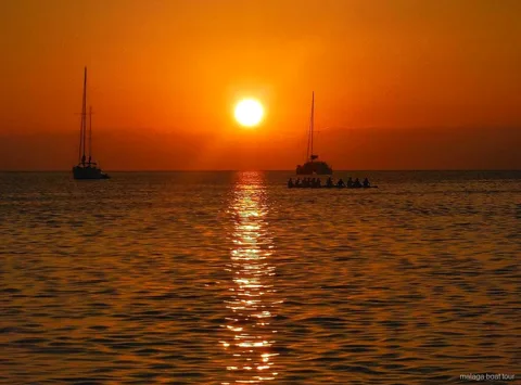 Atardecer naranja sobre el mar con dos veleros y un catamarán en silueta en un paseo en barco por Málaga
