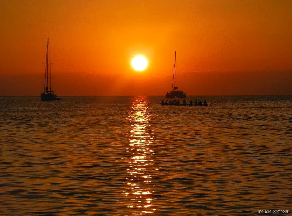 Atardecer naranja sobre el mar con dos veleros y un catamarán en silueta en un paseo en barco por Málaga