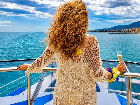 Vrouw in gehaakte jurk met een glas op het dek van een catamaran met zicht op de kust, boottocht Malaga
