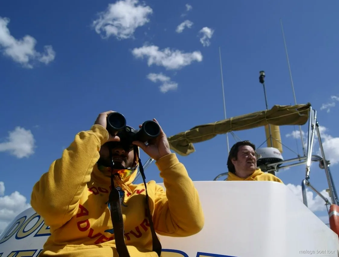 Tripulante con sudadera amarilla mirando con prismáticos bajo un cielo azul en una excursión en barco por Málaga
