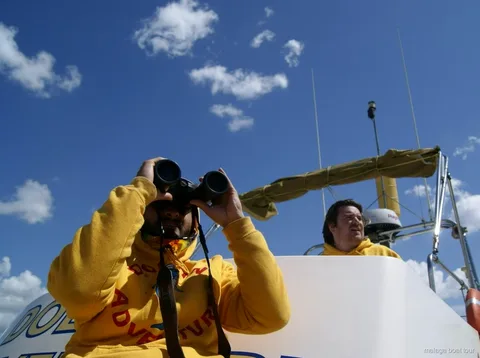 Crew member in a yellow sweatshirt scanning with binoculars under a blue sky on a Malaga boat tour